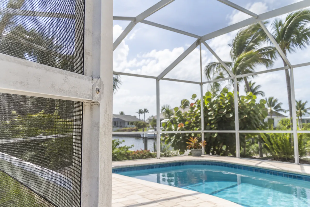 White coastal pool enclosure with lightly weathered frame hardware and palms beyond